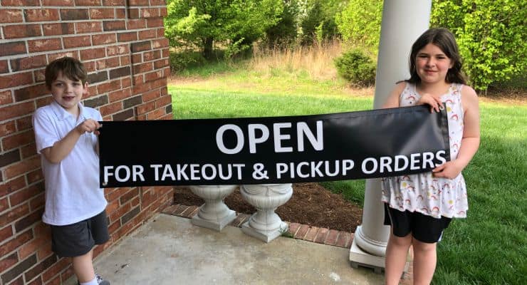 Two children hold a black sign with white lettering "Open for takeout and pickup orders"