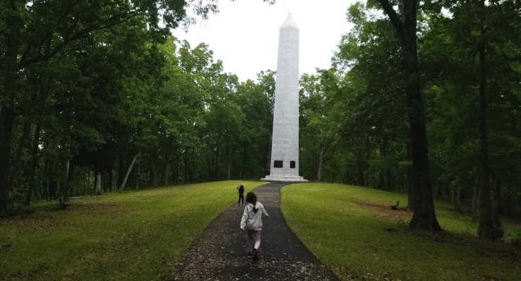 Two children running towards a tall white monument surrounded by trees.