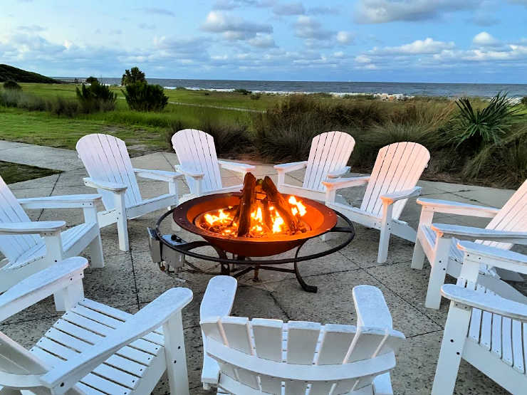 White Adirondack chairs surrounding a metal fire pit near the beach.