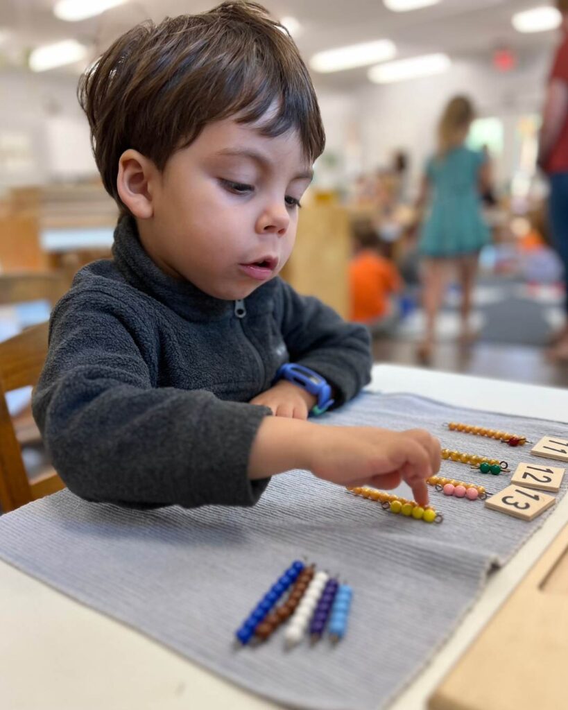 A young child concentrates while using colorful bead chains to count and match numbers on a classroom work mat.