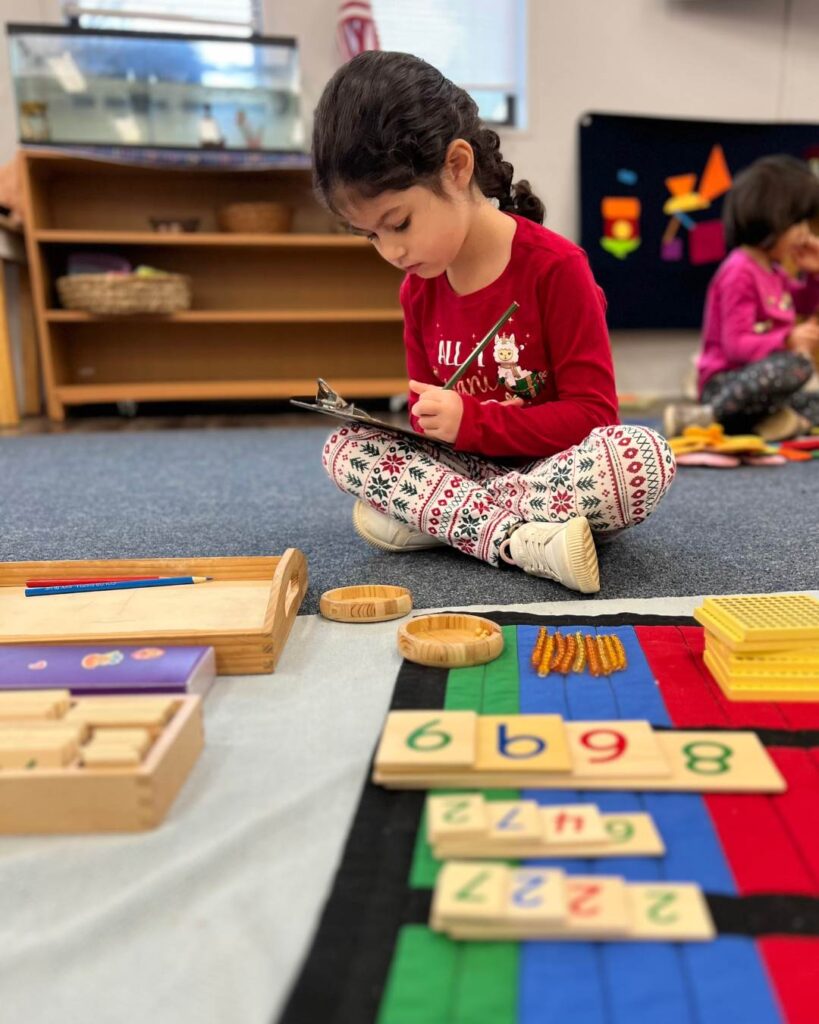 A child sits cross-legged on the classroom floor working independently with math materials and a clipboard, focused on completing a task.