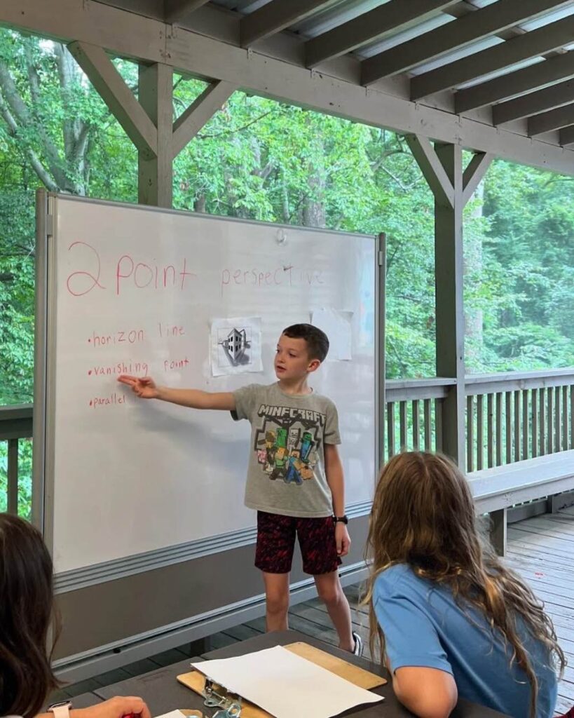 A student stands at a whiteboard explaining a two-point perspective lesson to classmates during an outdoor classroom session.