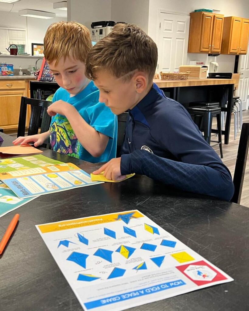 Two children sit at a table reviewing an instructional worksheet together, independently following steps and discussing their work.