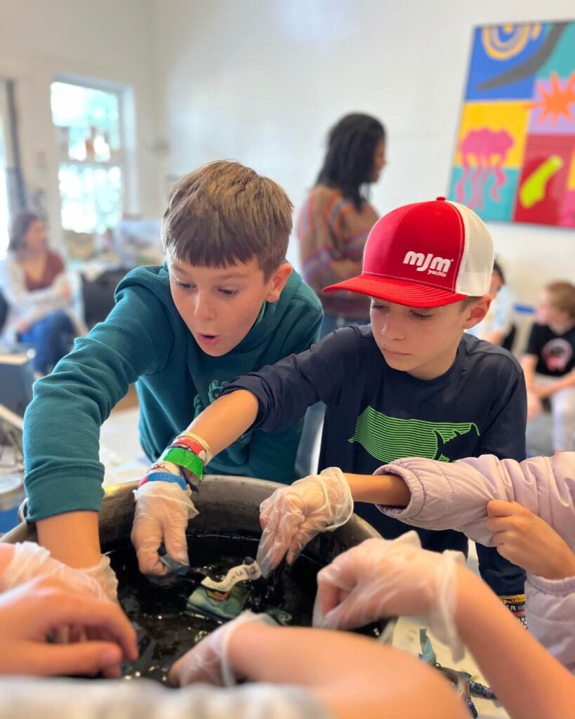 Children wearing gloves work together around a container during a hands-on science activity, exploring materials and observing results.