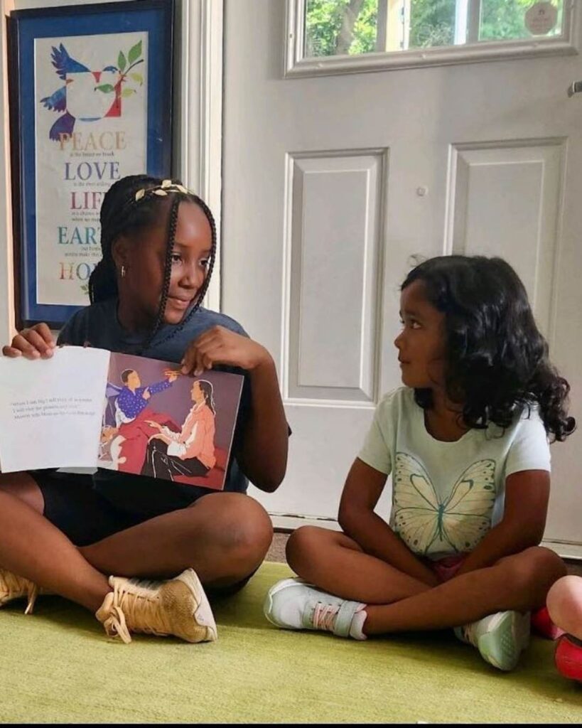 One child holds an open book while reading aloud to another child seated nearby, engaging in shared storytelling and listening.