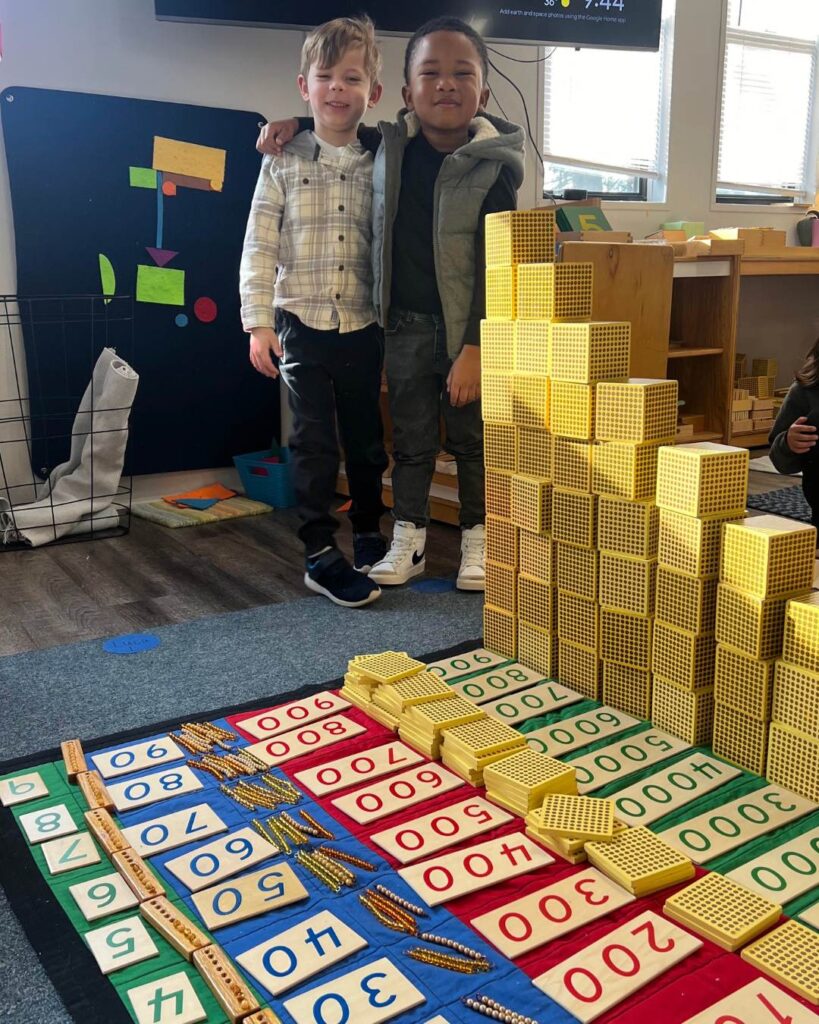 Two children stand beside a large math work on the floor made with number cards and base-ten blocks, smiling proudly after completing the activity.