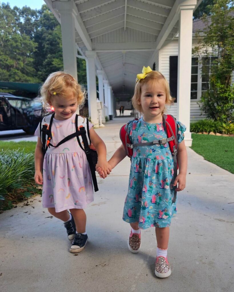 Two young children wearing backpacks walk hand-in-hand along a covered walkway outside a school building.