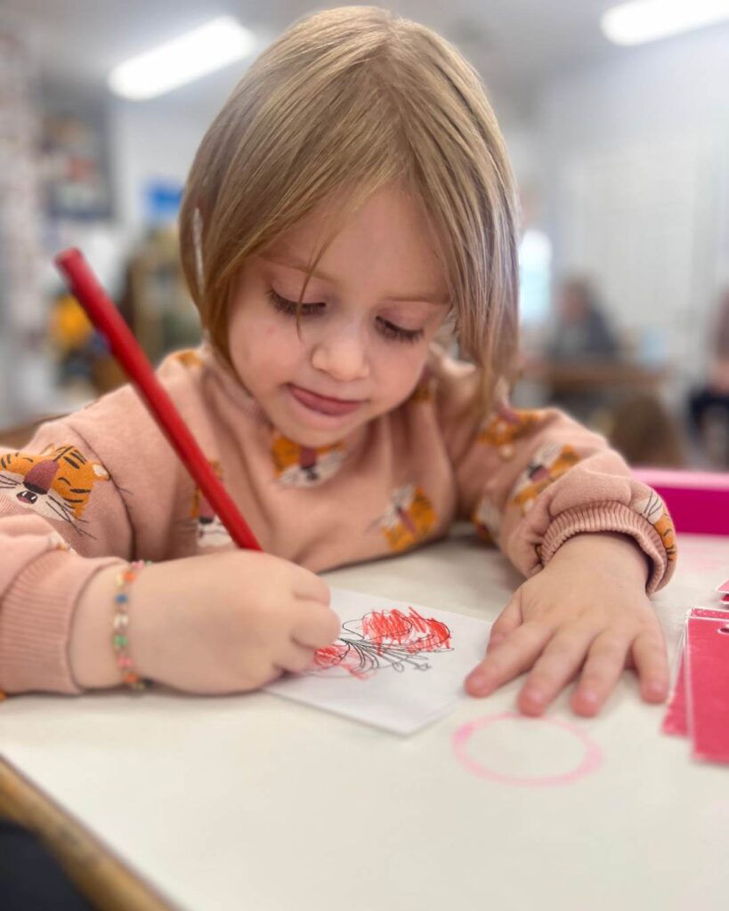 A child focuses on drawing with a red colored pencil at a classroom table, practicing fine motor skills.