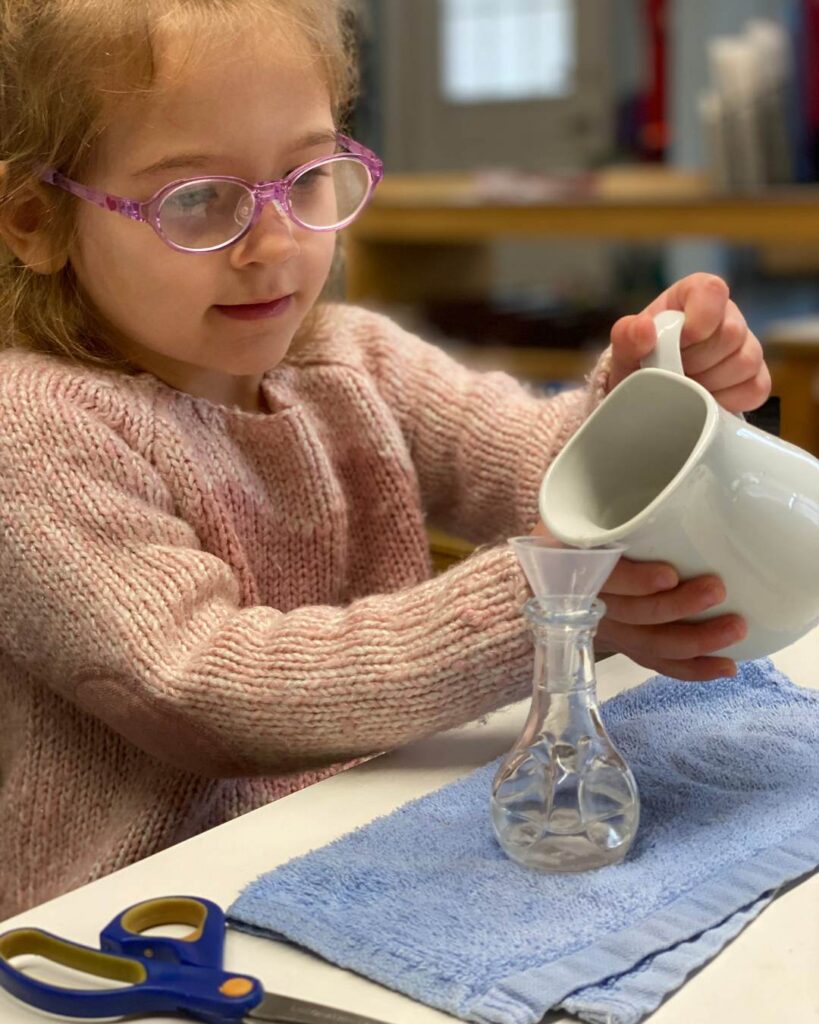 A child pours water from a pitcher into a small glass using a funnel, practicing focus, control, and independent task completion.