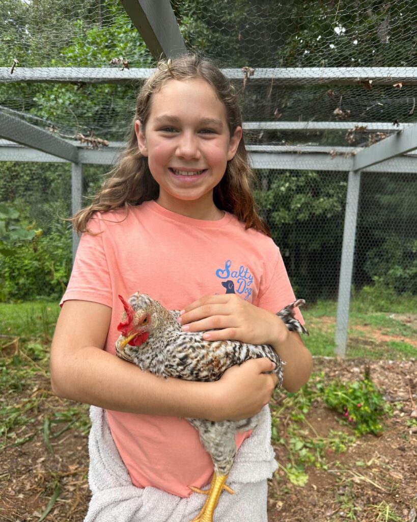 A smiling child gently holds a chicken outdoors, demonstrating responsibility and care for animals.