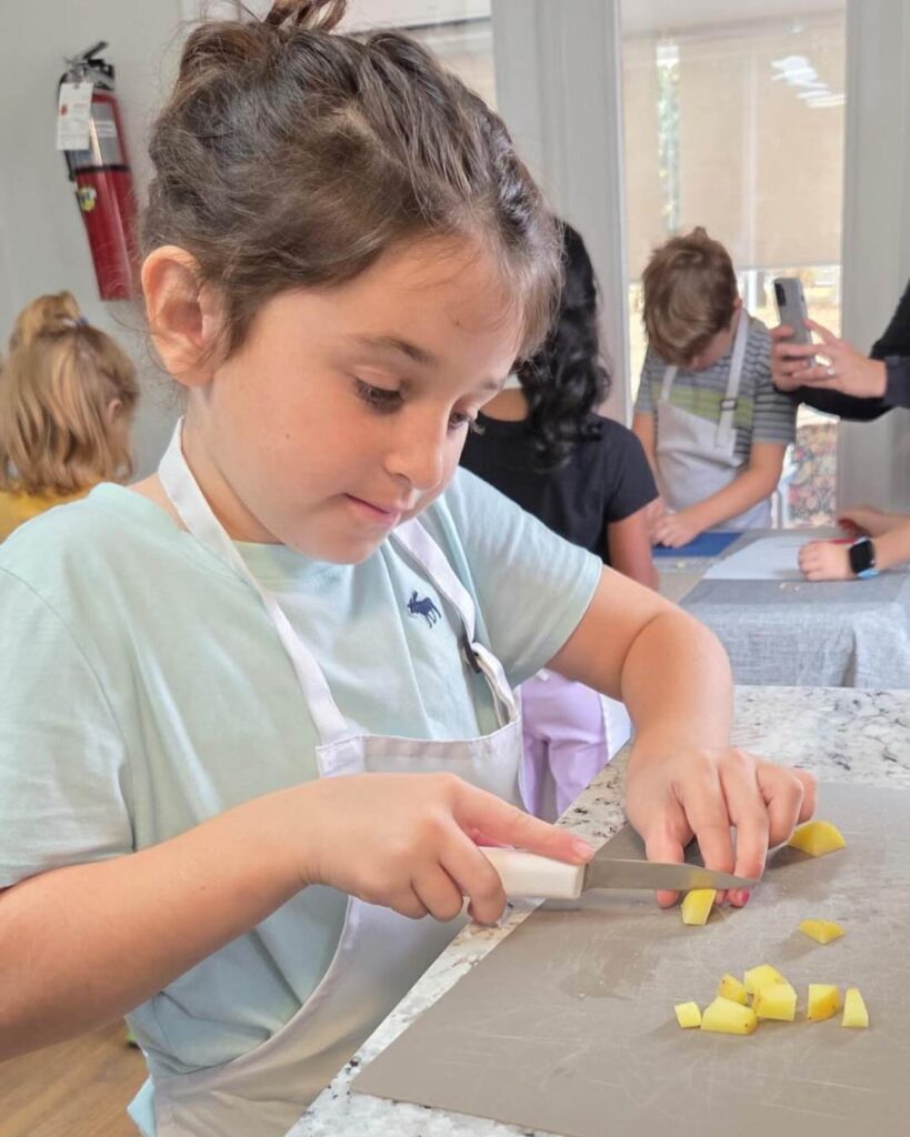 A child uses a child-safe knife to cut food on a work mat while wearing an apron, following steps independently during a classroom activity.