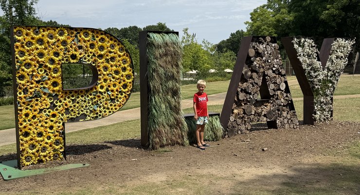 Child standing in front of the large PLAY sign made of sunflowers and natural materials at Gipson Play Plaza Dorothea Dix Park Raleigh NC