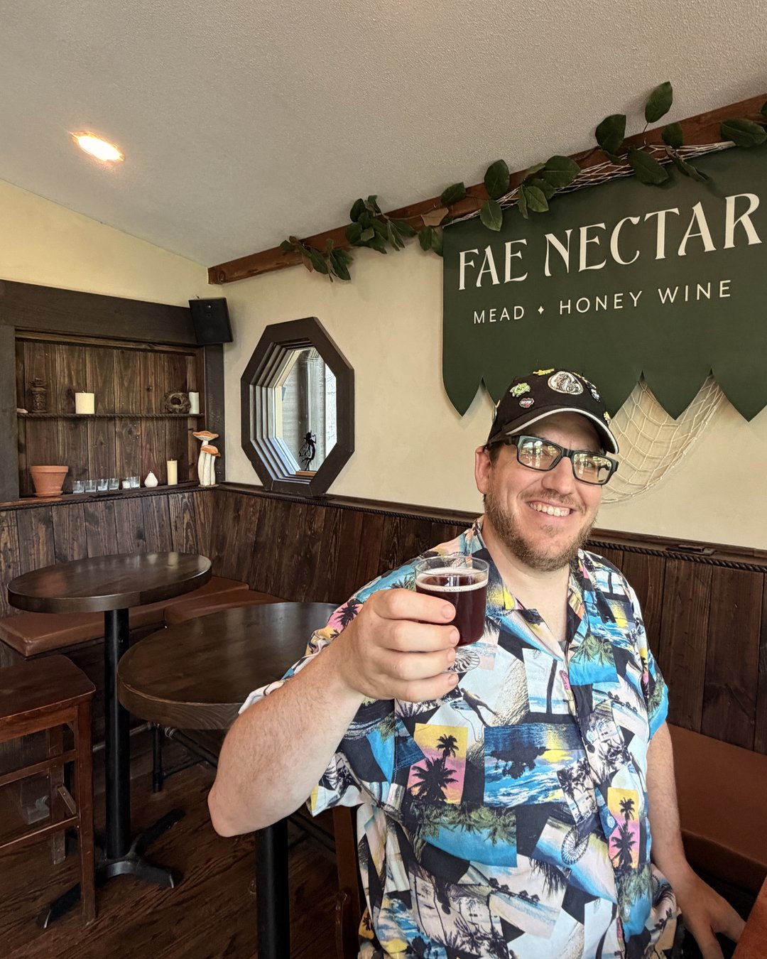 Man raising a glass of Elderberry Mead at Fae Nectar with the Fae Nectar Mead Honey Wine sign visible in background
