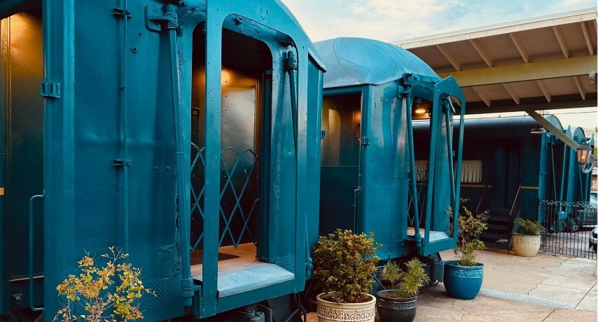Two blue train cars sit side by side in the fading sun with flowers by their steps