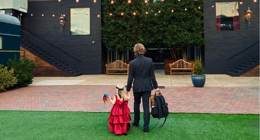 A father in a suit and young daughter in a dress hold hands and walk across the Arbor Lawn beneath string lights