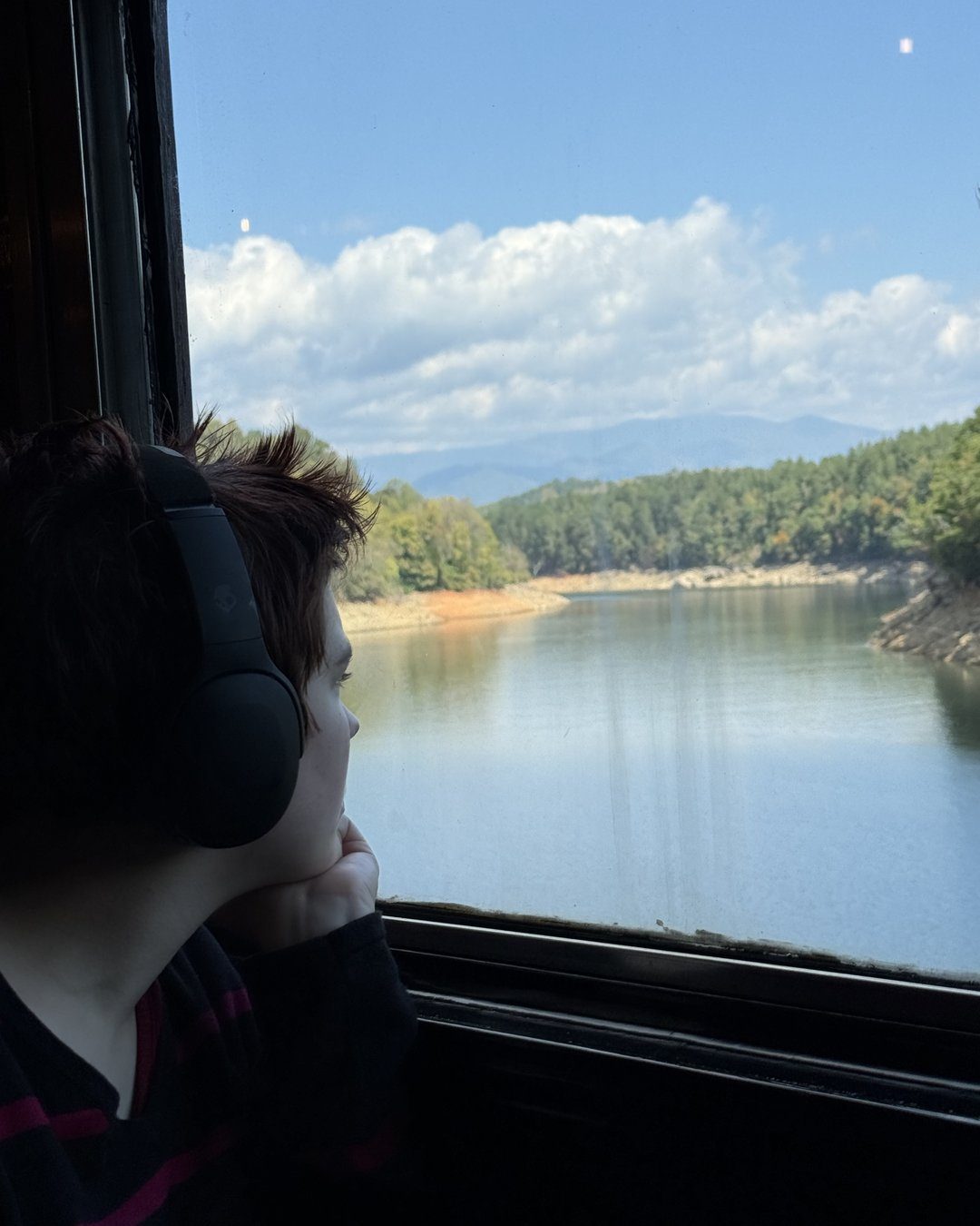 Teen wearing headphones looks out a train window at a calm lake with forested mountains beyond