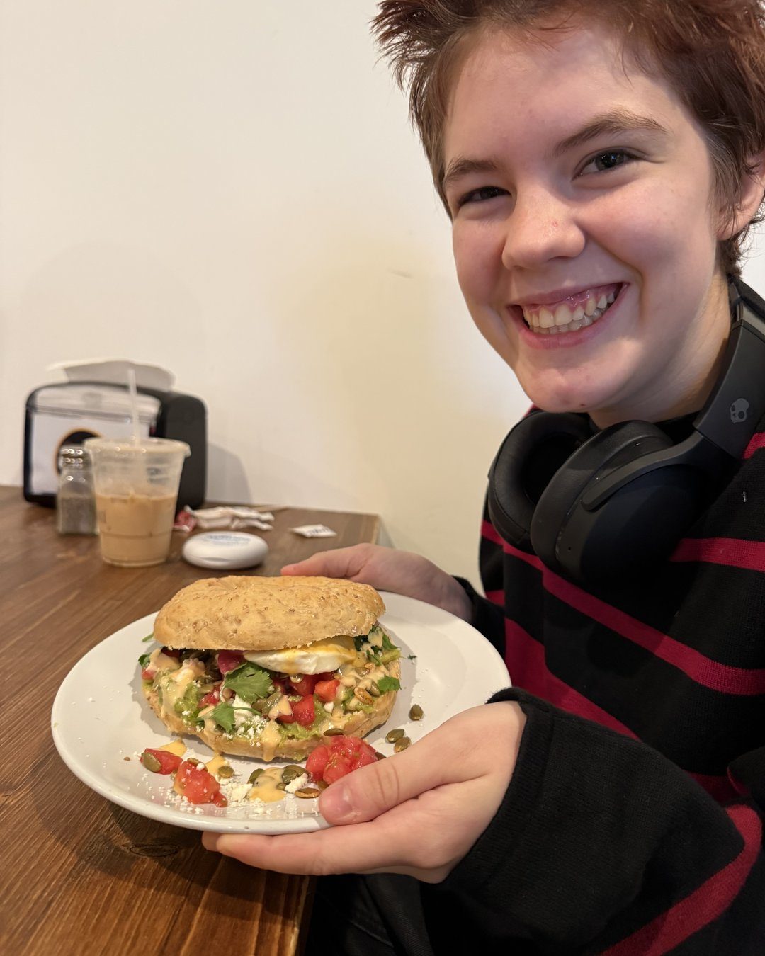 Smiling teen holding a plate with a breakfast bagel sandwich topped with egg, avocado and tomato